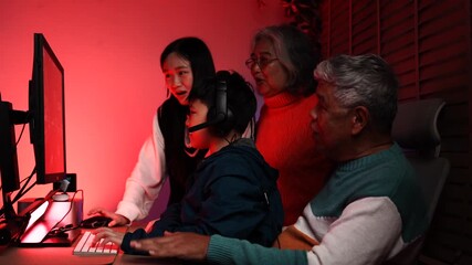 Asian family enjoys playing esports together at the monitor. Grandpa, Grandma and Granddaughter, Teenager and Grandson cheer on Grandpa playing online games at home. - Powered by Adobe