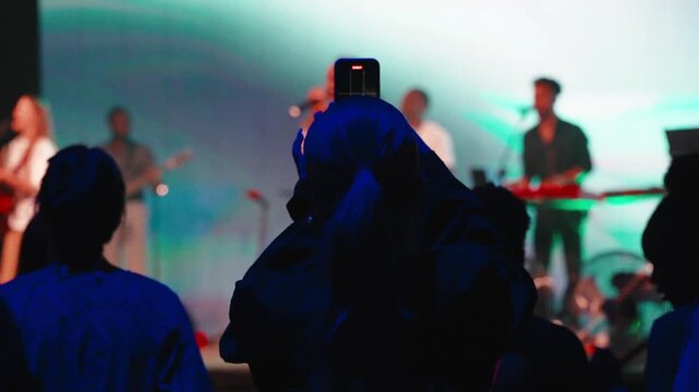 Dark-haired woman recording christian band performing onstage, bathed in colorful stage lighting during worship concert event