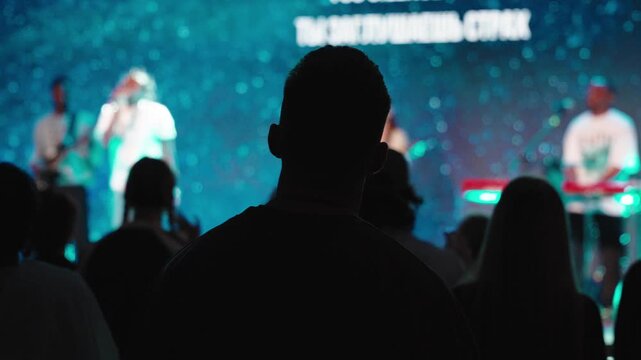 Silhouetted worshipper raising arms during christian concert, standing among crowd, band performing on stage with large screen displaying spiritual messages