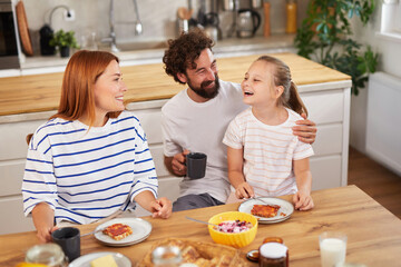 A happy family sits around a wooden table enjoying breakfast. The parents smile at their daughter while sharing a playful moment. The kitchen has a warm atmosphere with plants and natural light.