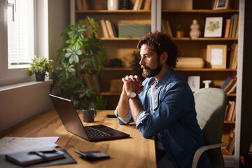 A man sits at a wooden desk in a home office, deep in thought while working on his laptop. Sunlight...