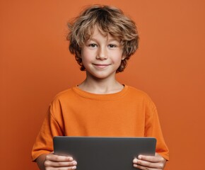A young boy, smiling and holding a tablet, stands against a vibrant orange background.