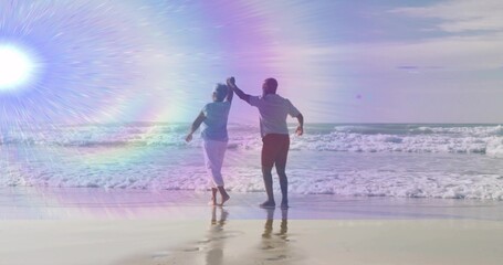 Dancing senior couple barefoot holding hands on beach shore, with breaking ocean waves, lens flare