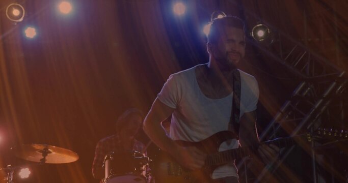 Strumming frontman playing electric guitar on stage under orange lights with drum kit and truss - Powered by Adobe