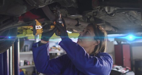 Working mechanic in blue coveralls using impact wrench under vehicle on hydraulic lift in workshop