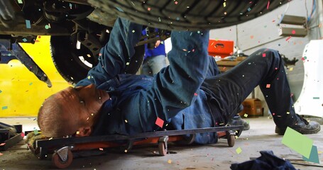 Mechanic lying on creeper inspecting suspension under raised vehicle at garage, drifting particles