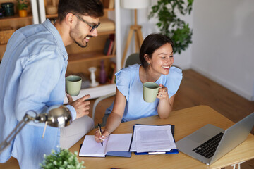 A couple engages in a productive work session at their home office, one taking notes while the other enjoys coffee, surrounded by a cozy atmosphere and greenery.