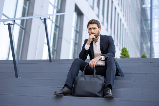 Pensive businessman in a suit is sitting on the steps outside a modern office building, briefcase at his side, deep in thought, contemplating his next move