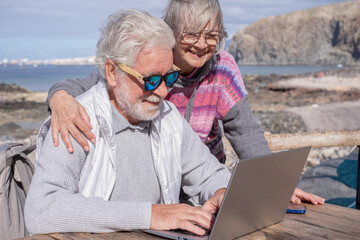Bonding senior couple using together laptop enjoying sunny day outdoor at the beach sitting at a wooden table Elderly people in vacation or retirement at sea