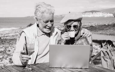 Black and white portrait of senior couple using together laptop enjoying sunny day outdoor at the beach having a break. Elderly people in vacation or retirement at sea