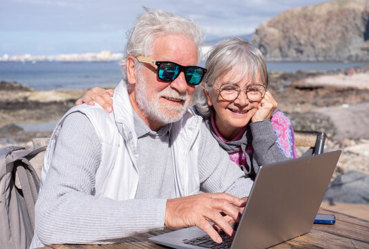 Bonding senior couple using together laptop enjoying sunny day outdoor at the beach sitting at a wooden table Elderly people in vacation or retirement at sea - Powered by Adobe