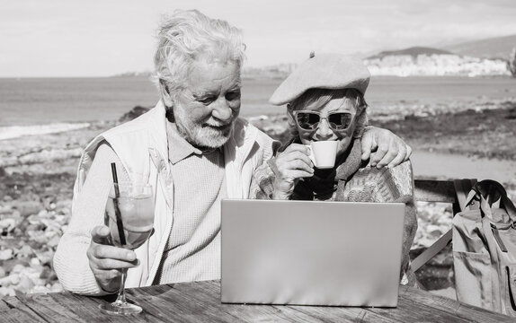 Black and white portrait of senior couple using together laptop enjoying sunny day outdoor at the beach having a break. Elderly people in vacation or retirement at sea