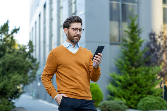 Business executive wearing glasses and an orange sweater, standing outdoors in a modern urban setting, uses a smartphone while holding one hand in his pocket - Powered by Adobe