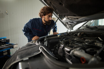 A skilled car mechanic inspects the engine under the hood of a vehicle in a repair shop. He carefully looks for any problems while ensuring all tools are available.