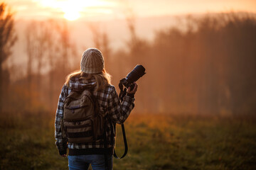 Woman photographer with backpack holding camera at sunset over autumn forest enjoying photographing nature in golden hour