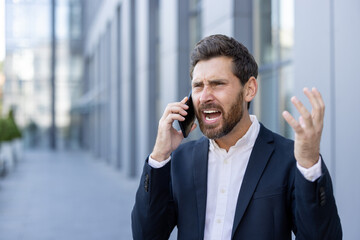 Furious businessman yelling into his smartphone while gesturing angrily, facing a stressful situation outside a modern office complex in the bustling city