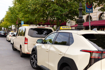Taxis sit patiently in a line on a city street, waiting for passengers. Trees provide shade, and buildings line the avenue, creating a bustling urban atmosphere