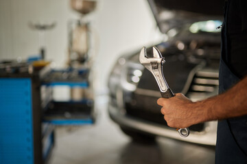 A mechanic holds an adjustable wrench, ready to fix a car in a busy garage. The environment is filled with various tools and equipment used for vehicle maintenance.