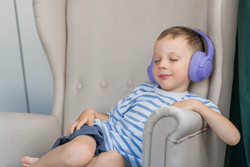 A little boy is sitting on the couch with purple headphones on. He is smiling and looking at the camera. He is wearing a striped T-shirt. Music for children's development.
