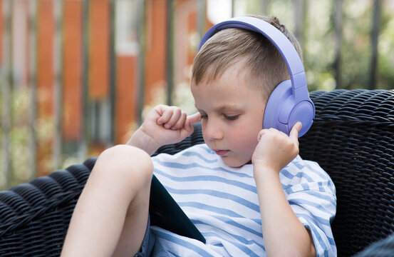 A 6-year-old boy is sitting on the couch with a smartphone in his hands and purple headphones on. The child is playing and listening to music. Children and gadgets.
