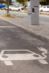 A clear marking for an electric vehicle charging station is visible on the pavement. The area is part of a city street, with parked cars and trees nearby, indicating a busy urban environment