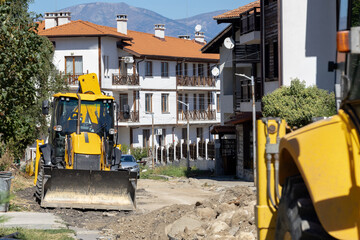 Heavy construction equipment, a yellow backhoe loader, is parked on a dirt road undergoing repairs, with modern apartment buildings and distant mountains under a clear sky.