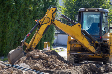 Obraz premium A yellow backhoe loader is actively digging a trench, surrounded by excavated earth and rocks, with green foliage in the background.