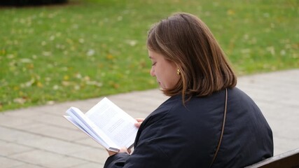 A young woman sits on a park bench reading a book, enjoying a peaceful autumn day surrounded by greenery and autumn fallen leaves. Horizontal 4k footage - Powered by Adobe