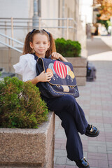Young girl in stylish outfit sits on stone wall with backpack in sunny city