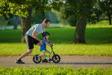 Young adult father teaching happy beautiful little boy to ride on first bike without pedals on sidewalk at city park. Learning to keep balance. Warm summer day. Cute 3 years old toddler. Side view.