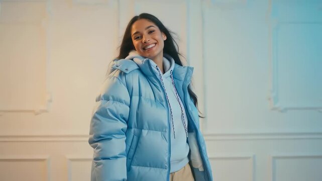 Cheerful young woman wearing a stylish blue padded jacket and hoodie, smiling confidently in a softly lit modern setting
