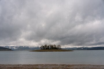 Fototapeta premium Moody autumn view of Batak Reservoir in Rhodope Mountains, Bulgaria, with small island under gray cloudy sky, scenic spot for eco tourism and nature travel