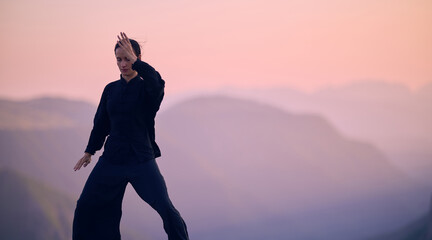 Woman practicing Tai Chi in traditional black outfit on mountain at sunrise, arms outstretched in...
