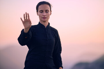 Fototapeta premium Woman practicing Tai Chi in traditional black outfit on mountain at sunrise, arms outstretched in meditation pose focusing on balance, energy, and mindfulness in nature.