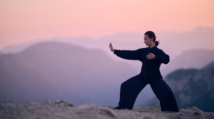 Woman practicing Tai Chi in traditional black outfit on mountain at sunrise, arms outstretched in meditation pose focusing on balance, energy, and mindfulness in nature.