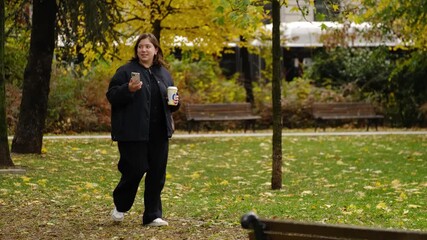 A young woman walks in city park with a take away paper coffee cup, using her smartphone for a video call on calm autumn day. Horizontal 4k footage