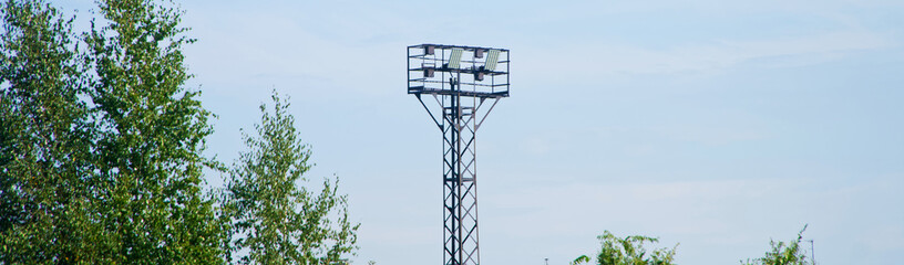 Tall metal tower with LED floodlights stands above green summer trees against blue sky, symbolizing modern street lighting. Photo