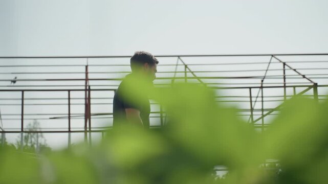 close up green leaves foreground with side view of agronomist walking past metal frame structure carrying crate and plier while rubbing nose during field inspection on sunny rural farm