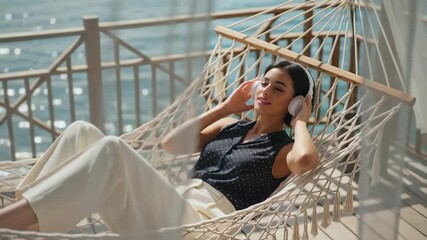 Young woman relaxing in a hammock by the seaside, wearing headphones and enjoying the sun - Powered by Adobe