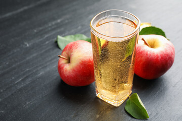 Fresh cider in glass, apples and green leaves on black table, closeup. Space for text