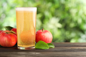 Delicious cider in glass, apples and green leaves on wooden table outdoors, space for text