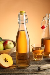 Delicious cider in bottles, glass and apples on wooden table against orange gradient background, closeup