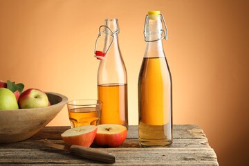 Delicious cider in bottles, glass, apples and knife on wooden table against orange gradient background