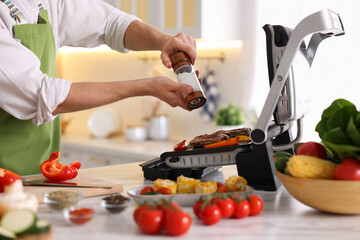 Man adding salt to vegetables and meat cooking on electric grill in kitchen, closeup