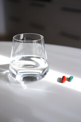 Transparent water glass and pills capsules on white surface. Daily vitamins and medicine.