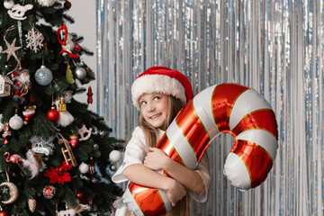 Cheerful 9-year-old girl in Santa Claus hat holding foil balloon shaped like candy cane in front of decorated Christmas tree, festive holiday mood