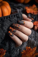Una mano femminile con unghie decorate con motivi di zucca color arancione su uno sfondo nero lucido, circondato da zucche e foglie autunnali per festeggiare  Halloween.