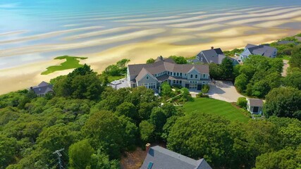Drone footage of a large luxury property with elegant design, landscaped gardens, and sweeping views of the ocean, sitting above a sandy shoreline and calm blue waters under clear skies.