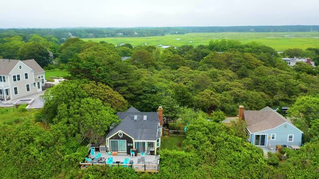 Scenic aerial view of a Cape Cod home with a large deck and outdoor seating, nestled among dense trees, overlooking expansive marshlands and tidal waterways in the distance.
