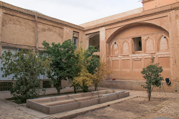 Obraz premium Traditional courtyard with trees and old adobe walls in Yazd, Iran, showcasing Persian architecture and desert lifestyle.